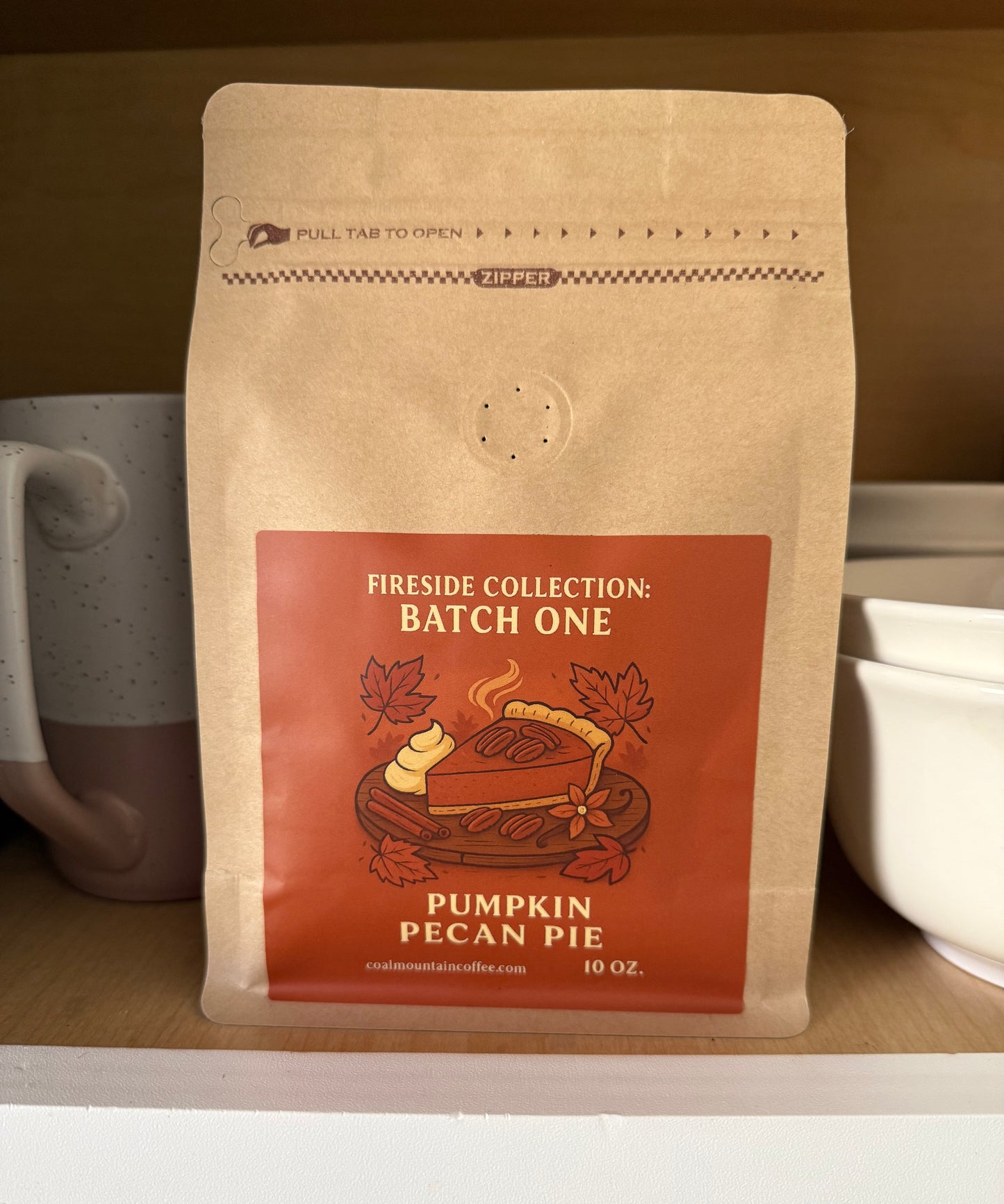 Brown coffee bag labeled 'Pumpkin Pecan Pie' from the 'Fireside Collection: Batch One' on a wooden shelf with mugs.